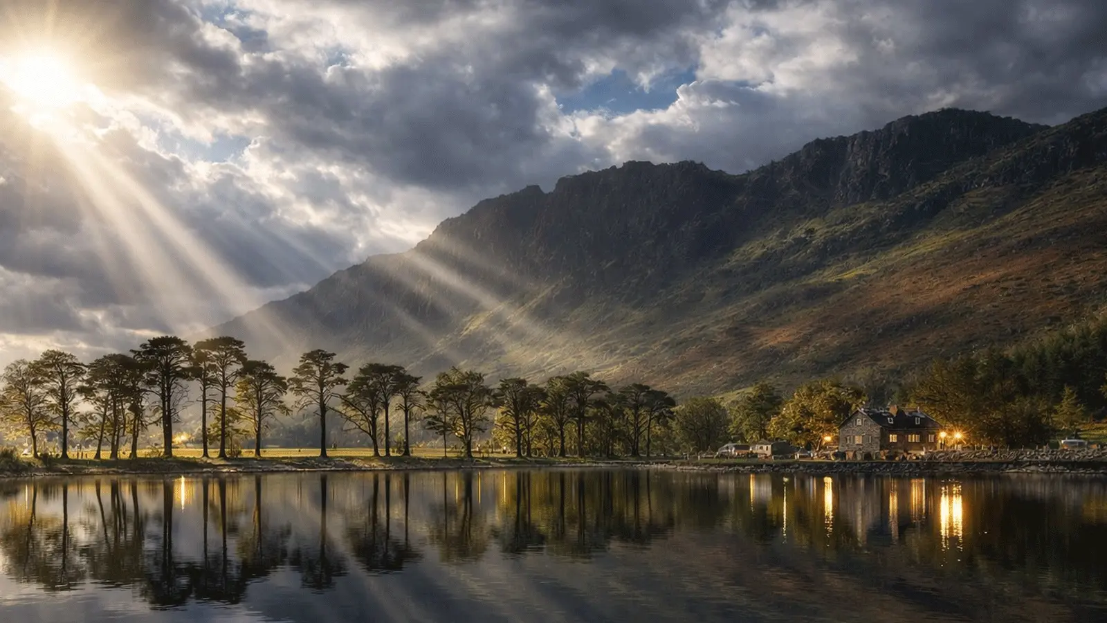 Sun rays over a Lake District mountain and calm lake, with trees and cottages reflected in the water